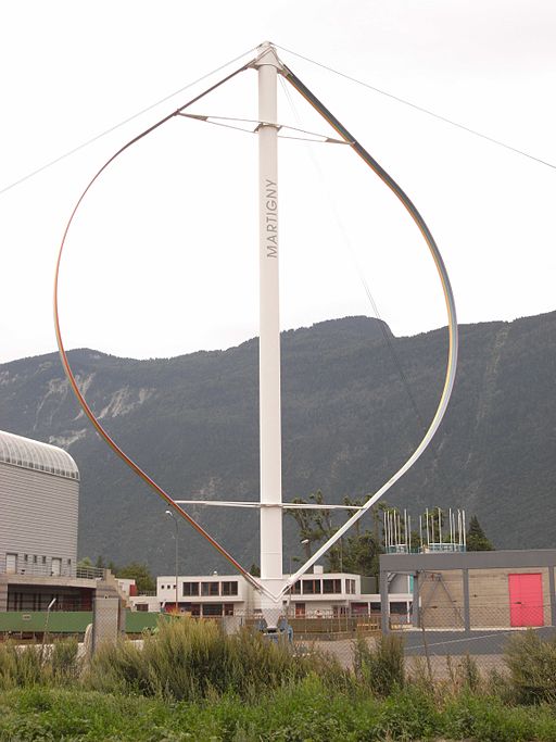 A vertical-axis wind turbine with curved blades mounted on a central tall white tower labeled "MARTIGNY." The turbine is located outdoors with mountains in the background and several buildings, including industrial and residential structures, in the foreground. The design shows symmetrical blades extending from the top and bottom of the central shaft, forming a teardrop-like shape.